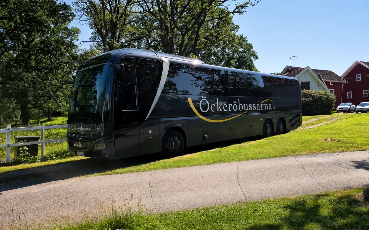 Öckeröbussarnas turistbuss under stora lövträd, med röda trähus i bakgrunden